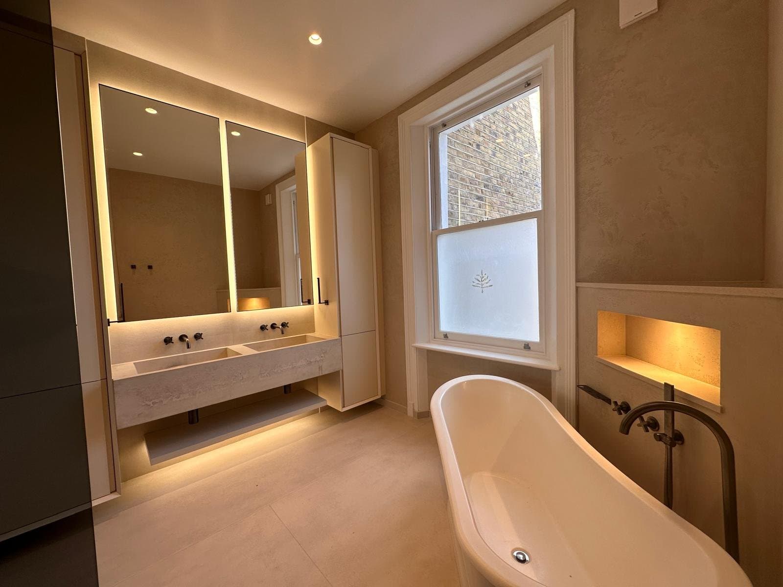 Bathroom with a freestanding oval bath and a double vanity with backlit mirrors, in a period property with a sash window