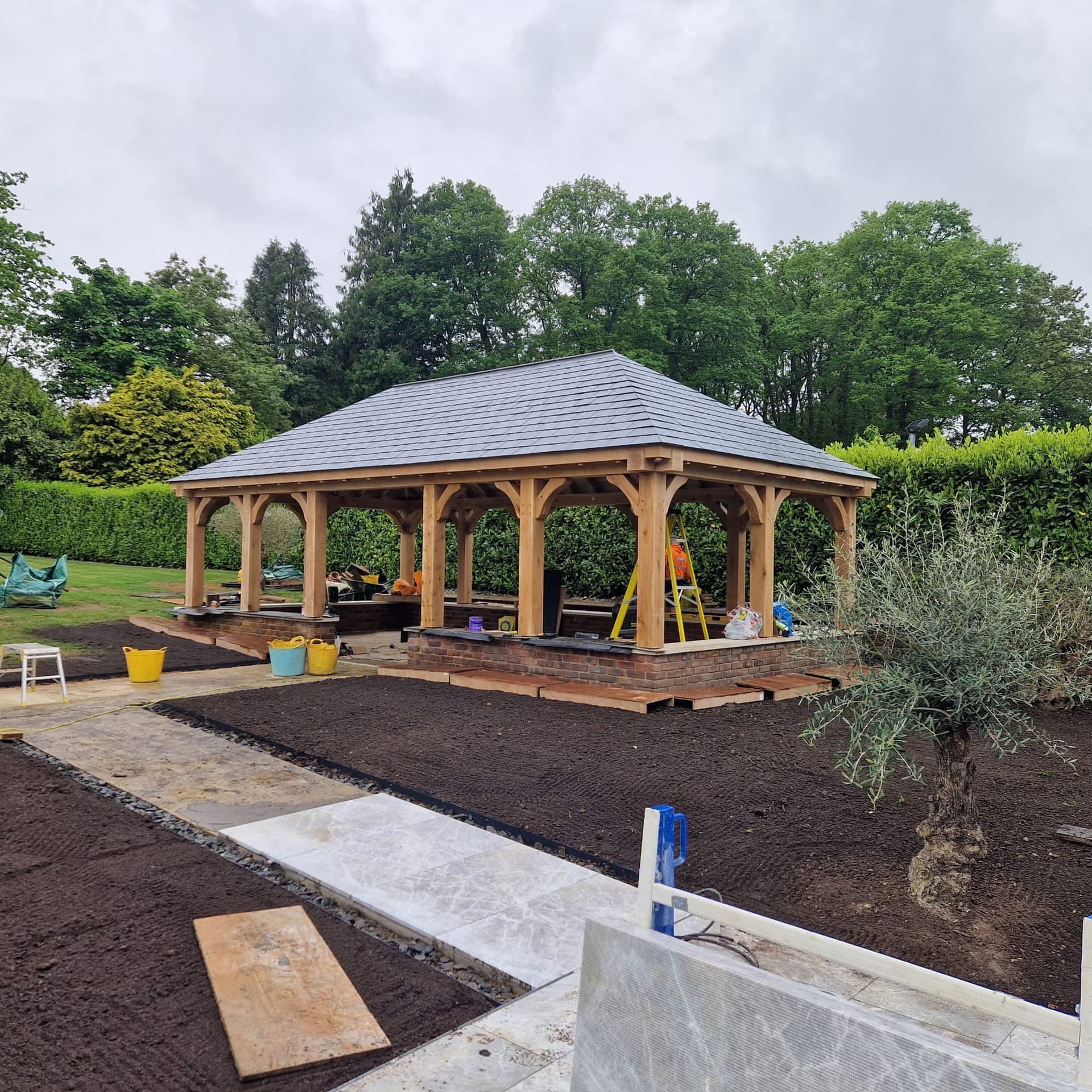 Green oak-framed barbecue pavilion with a hipped slate roof in a landscaped garden